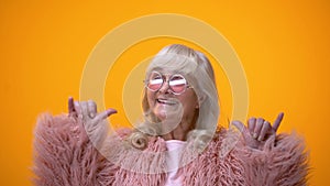 Funny positive elderly female in pink coat making rocker gesture, having fun