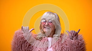 Funny positive elderly female in pink coat making rocker gesture, having fun