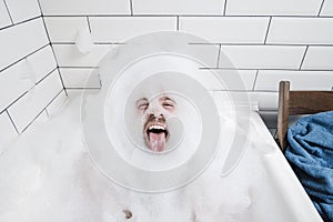 Funny, positive man lies in a bathtub with lush foam and shows his tongue.