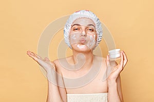 Funny positive woman with pout lips, applies moisturising facial mask, holding nutritious cream in hands, wears shower cap sending