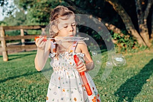 Funny preschool Caucasian child girl blowing soap bubbles in park at summer sunset