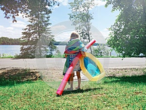Funny preschool girl with many beach towels and swimming noodle standing near lake river ready to swim on summer day