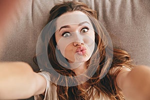 Funny pretty lady lies on sofa indoors make selfie.