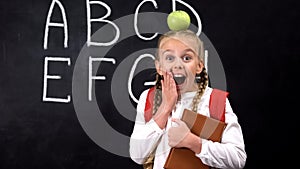 Funny pupil with apple on head standing against blackboard, education concept