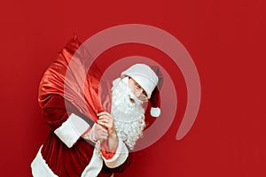 Funny Santa Claus carries on his back a bag with gifts isolated on red background. Portrait of Santa carrying presents to children