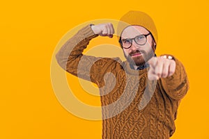 Funny serious hipster caucasian man with a beard, glasses, and a beanie standing over yellow background pointing at