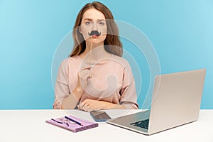 Funny serious woman office employee sitting at workplace, covering lips with fake paper moustache, imitating strict boss