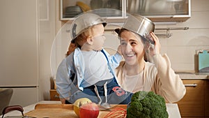 Funny shot of little baby boy with bowl on head looking at mothre with cooking pan on kitchen