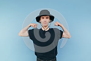 Funny skinny young man in panama and black t-shirt shows biceps to camera and smiling, isolated on blue background