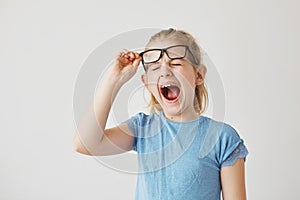 Funny small blonde girl with pretty blue eyes raising glasses and yawning with closed eyes in classroom because long