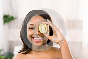 Funny smiling african american young woman holding kiwi half in front of her eye over white background