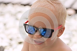 Funny smiling baby boy in sunglasses on the beach. Stones are reflected in the glasses. Little boss on vacation