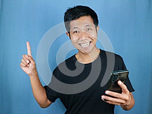 Funny smiling face expression asian man smile holding mobile phone with gesture pointing finger up photo
