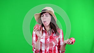 Funny, smiling female farmer in checkered shirt and hat, holding in hands two large sour apples, eats, bites them