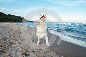 Funny smiling laughing white Caucasian child kid baby girl in fur coat and tutu skirt running on ocean sea beach on sunset