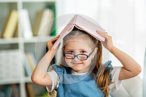 Funny smiling little girl with glasses and book on her head is sitting at table