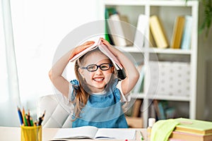 Funny smiling little girl with glasses and a book on her head sitting at table