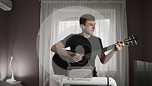 Funny smiling man is playing guitar while walking on treadmill at home. Fit athletic male walks on running machine and plays on gu