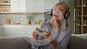 Funny smiling mature caucasian woman playing video game console in headphones using joystick controller at home on couch