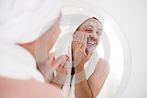 Funny smiling young man applying facial cream looking at the mirror. S