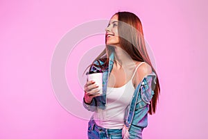 Funny stylish young woman in trendy blue denim jacket in vintage white t-shirt with cup of tasty drink posing near pink wall in