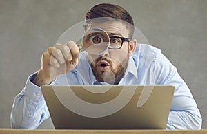 Funny surprised businessman with laptop looking at something through magnifying glass