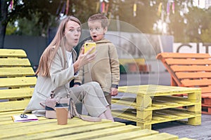 Funny and surprised young mom with preschooler son sit on bench using smartphone and have fun together. Mother and little boy