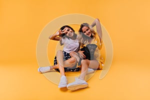 Funny tanned girls posing on the floor with dog. Studio shot of stunning white sisters isolated on yellow background