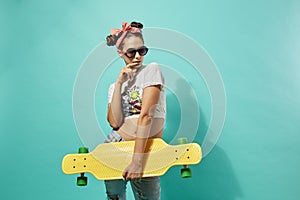 Funny teen girl in sunglasses and pink bow on her head dressed in jeans and top stands with yellow skateboard on the