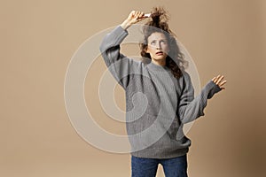 Funny thoughtful curly beautiful female in gray casual sweater with hairbrush comb pulls curls up posing  on