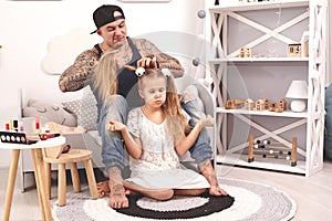 Funny time Tattoed father in a cap and his child are playing at home. Dad is doing his daughter`s hair in her bedroom