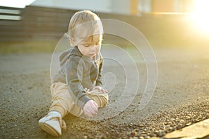 Funny toddler boy sitting on the ground outdoors on sunny summer day. Child exploring nature