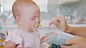 Funny toddler sitting in baby chair and crying, refusing to eat during feeding with spoon by mom.