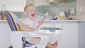 Funny toddler sitting in baby chair and crying, refusing to eat during feeding with spoon by mom.