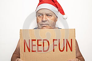 Funny upset and angry mature man in red Christmas Santa hat holds cardboard sign with inscription - need fun on white background