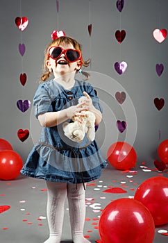 funny white Caucasian little girl toddler in studio with red balloons hearts on grey background wearing funny glasses