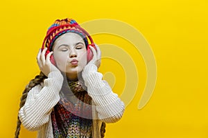 Funny Winsome and Smiling Caucasian Blond Girl In Warm Knitted Hat and Scarf While Listening to Music in Wireless Headphones Over