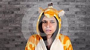 A funny woman in a big pajamas of giraffe is showing a fright on a brick background. A brunette was gripped by fear.