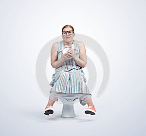 A funny woman in glasses and a colored dress is sitting on the toilet holding toilet paper in her hands, on a light blue