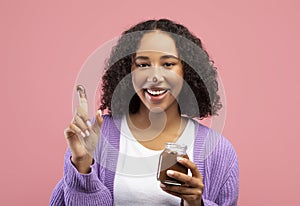Funny young African American woman eating chocolate paste from jar with finger, having dirty nose, enjoying dessert