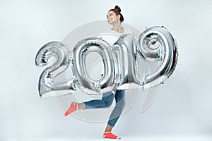 Funny young girl dressed in white t-shirt, jeans and pink socks holding balloons in the shape of numbers 2019 on the