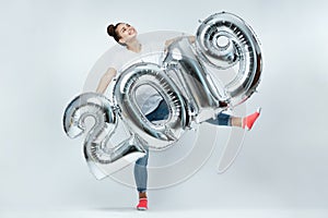 Funny young girl dressed in white t-shirt, jeans and pink socks holding balloons in the shape of numbers 2019 on the