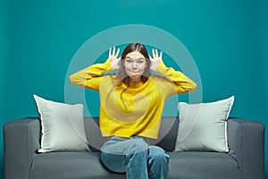 Funny young girl making faces, grimacing, fooling, showing big ears by hands, teasing, sitting on sofa