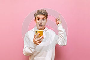 Funny young man with a disgruntled face stands on a pink background with a smartphone in his hands, looks into the camera and