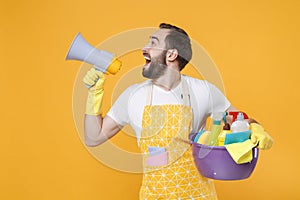 Funny young man househusband in apron rubber gloves hold basin with detergent bottles washing cleansers doing housework