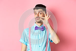 Funny young man touching his french moustache and pucker lips, looking silly at camera, standing in bow-tie and