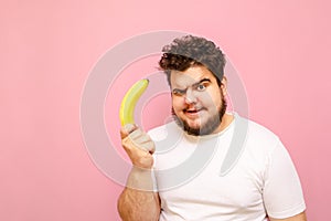 Funny young overweight man standing with a banana in his hand on a pink background, looking into the camera and making a funny