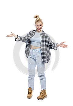 A funny young teenage girl stands with her arms spread to the sides. Laughing blonde girl in jeans, blue top and plaid shirt.