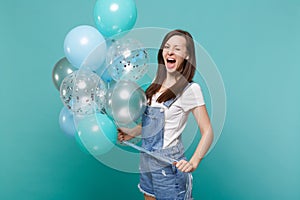 Funny young woman in denim clothes blinking, keeping mouth wide open, celebrating, holding colorful air balloons