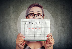 Funny young woman in glasses hiding behind a periods calendar and looking at camera